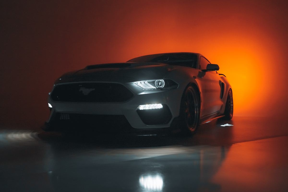 Gray Ford Mustang with headlights on against an orange-lit studio backdrop