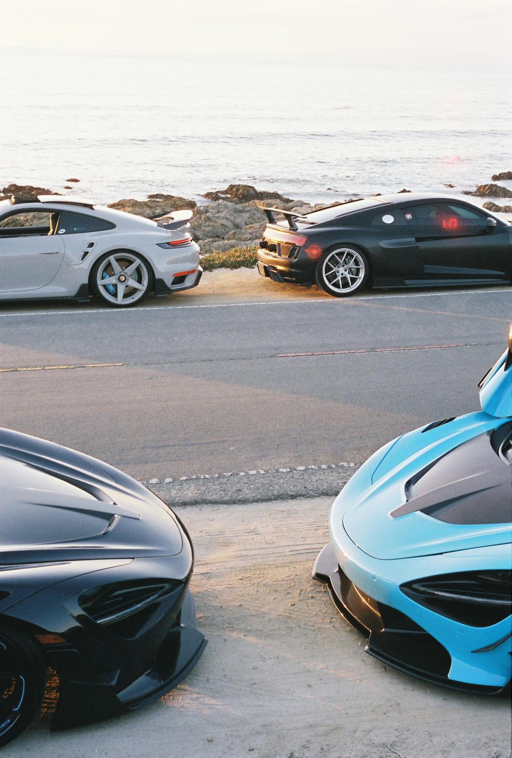 Two McLaren noses in the foreground with a Porsche 911 and Audi R8 parked along the ocean